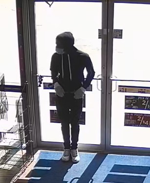 Camera photo of another suspect standing at the door. Suspect is wearing black hoodie and pants, white sneakers, gray gloves and a cloth mask.